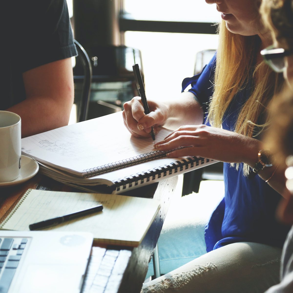 A woman in a blue top writes in a spiral notebook while collaborating with colleagues around a table. Coffee cups, laptops, and additional notebooks are spread across the workspace, showing an active design planning session.