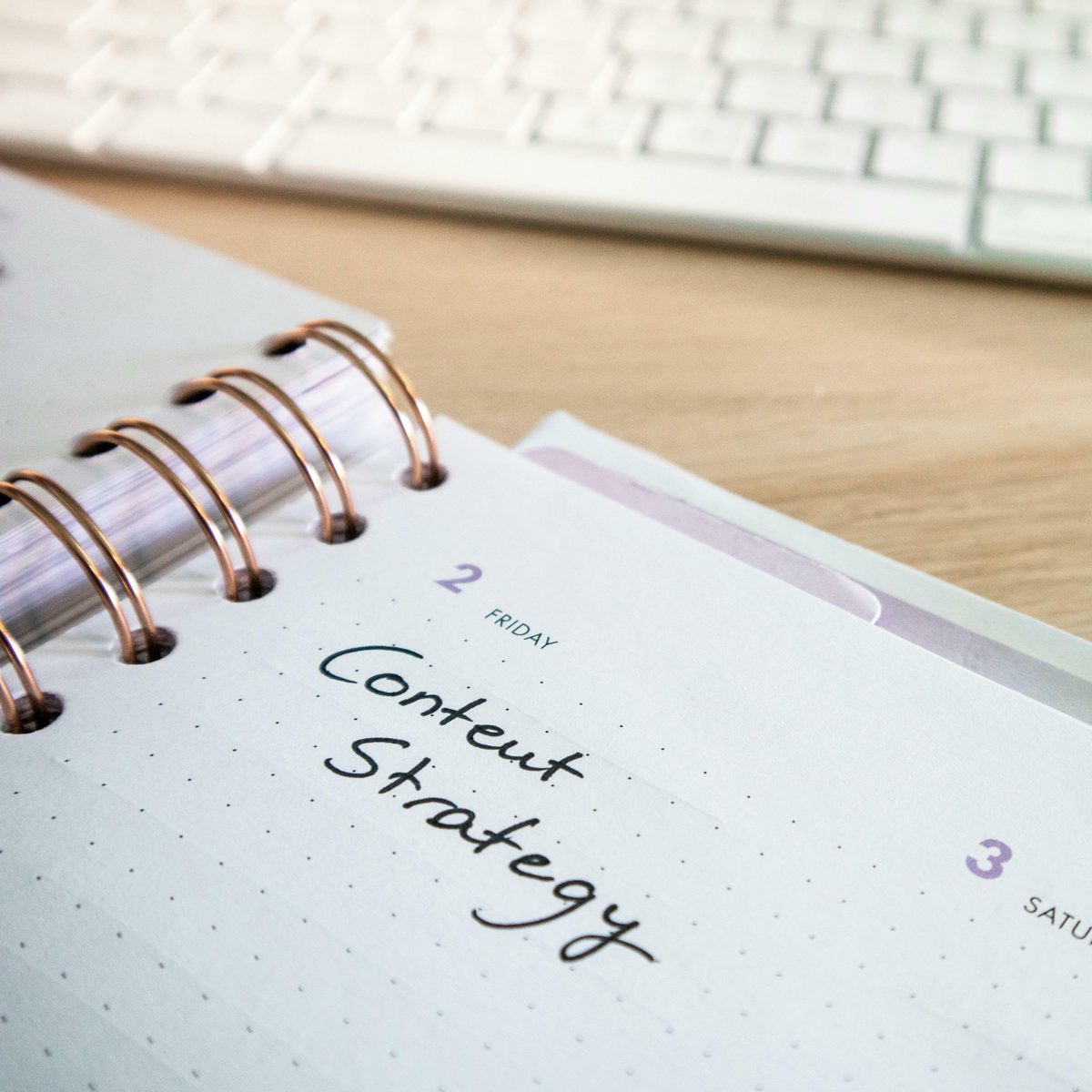 Close-up of an open spiral-bound planner showing 'Content Strategy' handwritten on Friday's page, with a keyboard visible in the background on a wooden desk